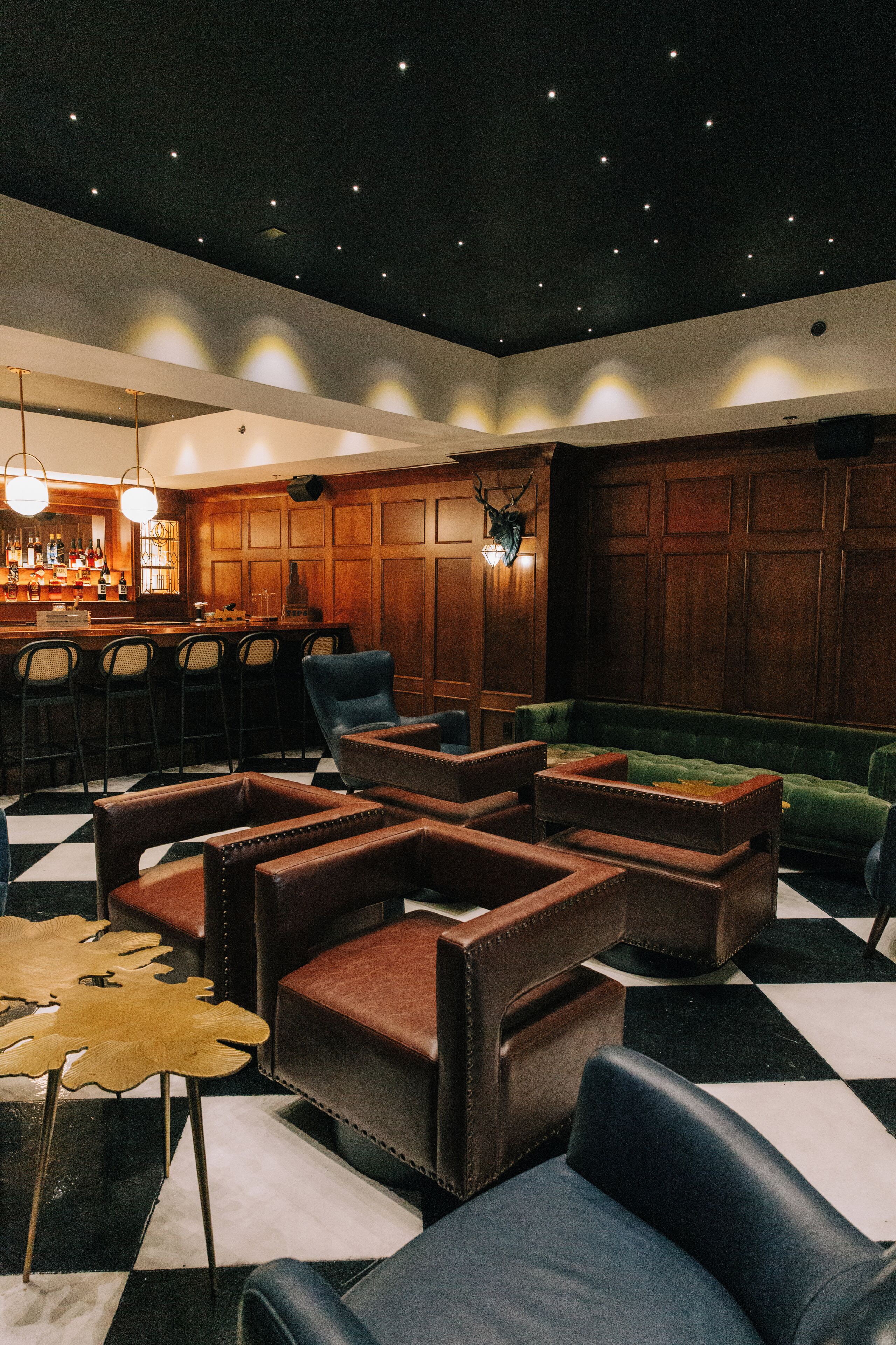 Uma área de bar com banquetas, balcão com garrafas e um espaço de estar com sofás de couro e piso xadrez.