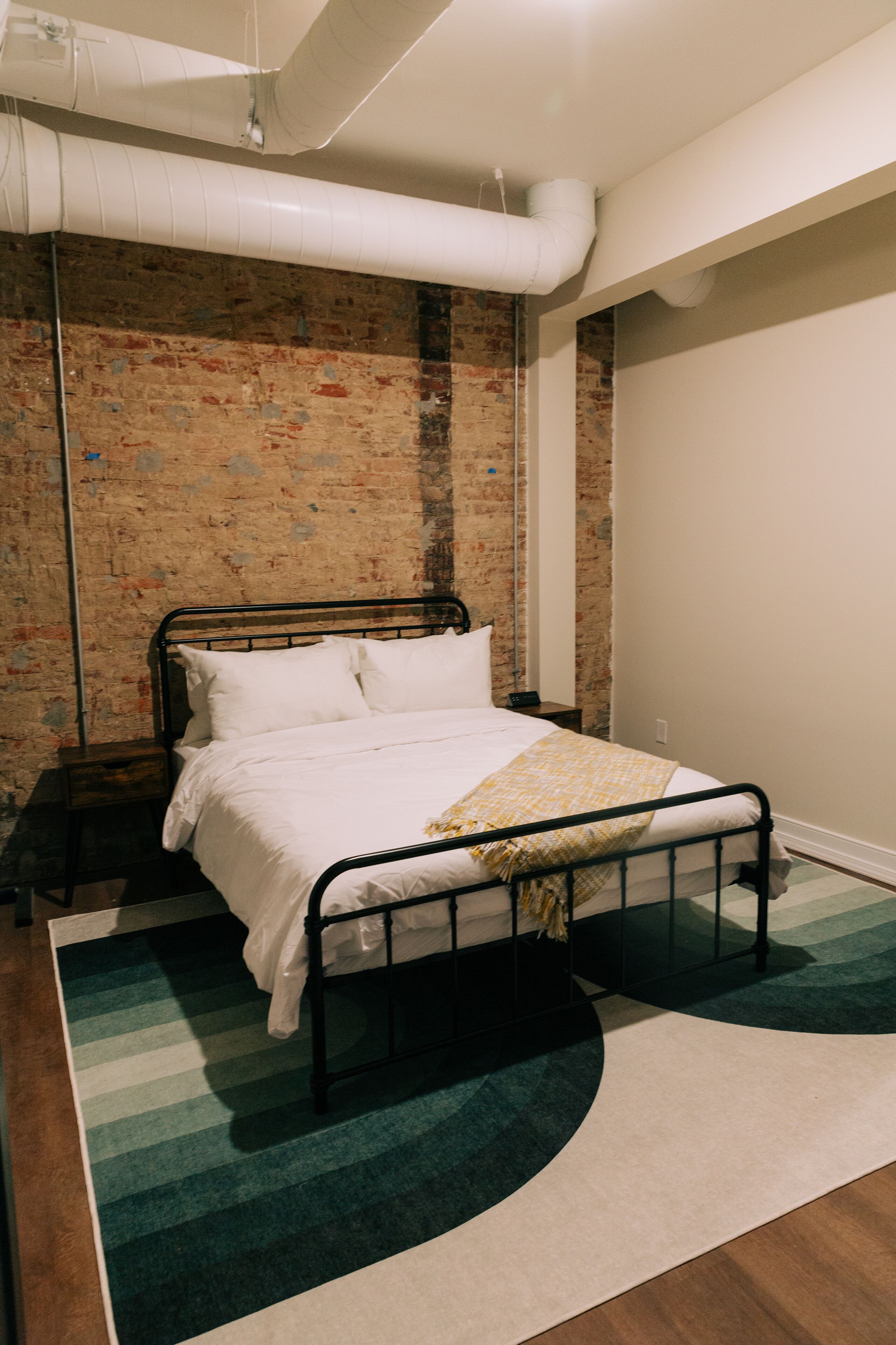Un dormitorio con estructura de cama de metal, ropa de cama blanca, pared de ladrillo y alfombra verde y blanca.