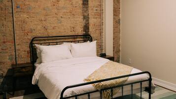 Un dormitorio con estructura de cama de metal, ropa de cama blanca, pared de ladrillo y alfombra verde y blanca.
