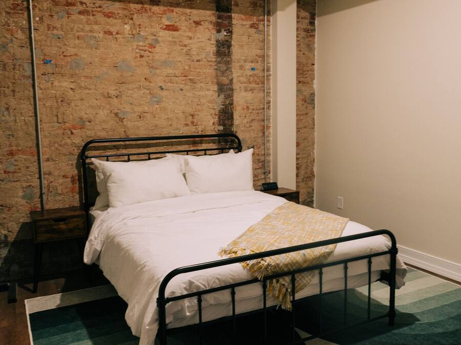Un dormitorio con estructura de cama de metal, ropa de cama blanca, pared de ladrillo y alfombra verde y blanca.