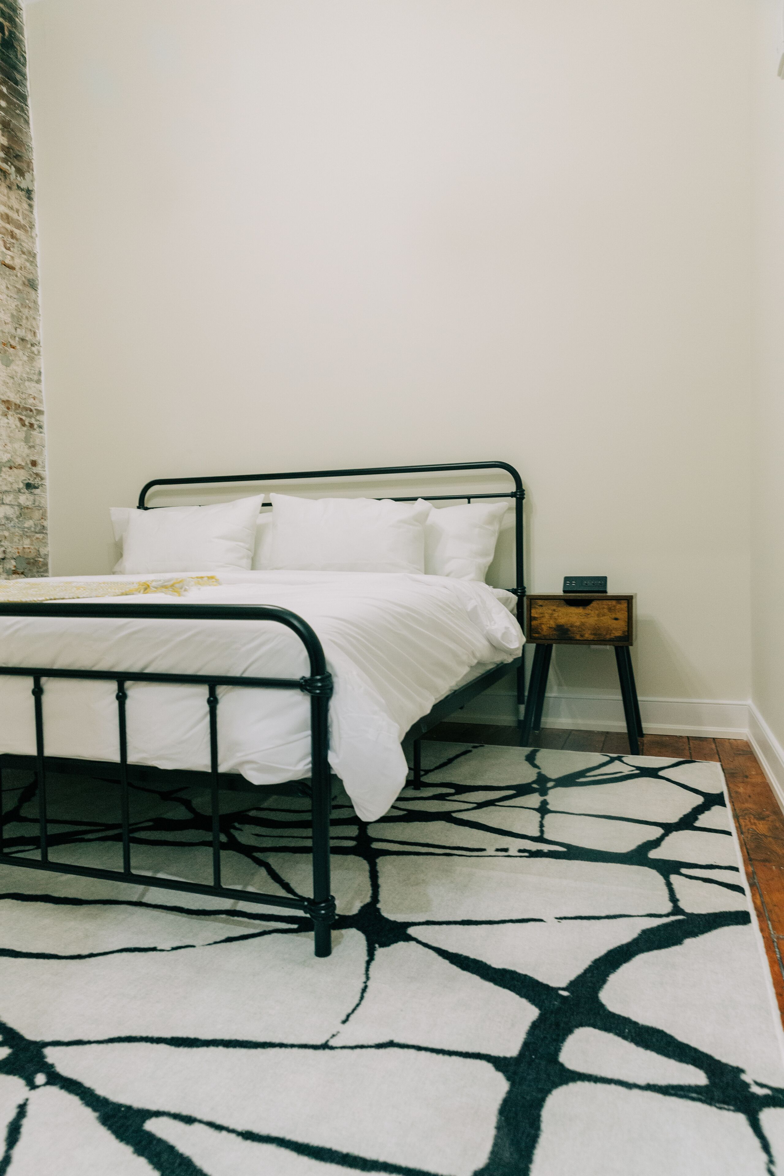 Una cama con ropa de cama blanca, estructura de metal negro y una mesita de noche de madera con un reloj.
