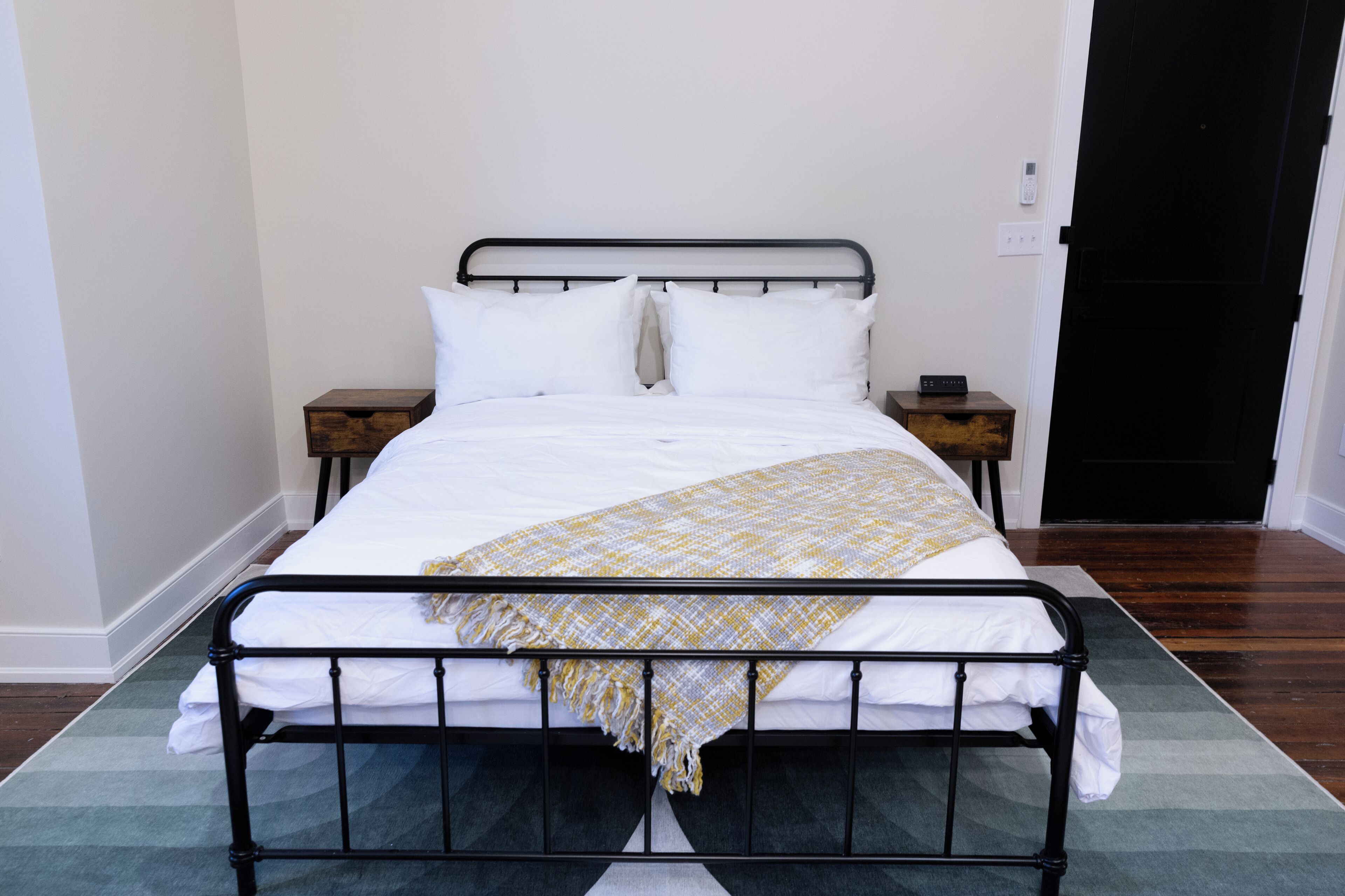 A neatly made bed with white linens and a patterned throw blanket, a bedside table with a clock, and a door with a black handle.