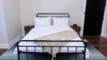 A neatly made bed with white linens and a patterned throw blanket, a bedside table with a clock, and a door with a black handle.