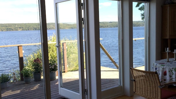 A room with large windows overlooking a lake, wooden deck, and a view of the forest.