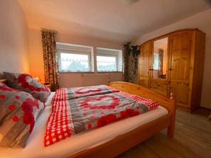 Ein Schlafzimmer mit einem hölzernen Bett, zwei Fenstern, einem Kleiderschrank und einer Pflanze.