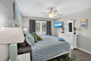 Un dormitorio con cama, ventilador de techo, cómoda, televisor y ventana con cortinas.