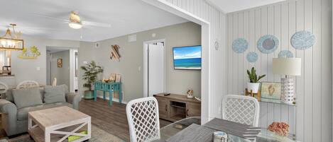 A modern living room with a glass dining table, white chairs, a grey sofa, and a TV mounted on the wall.
