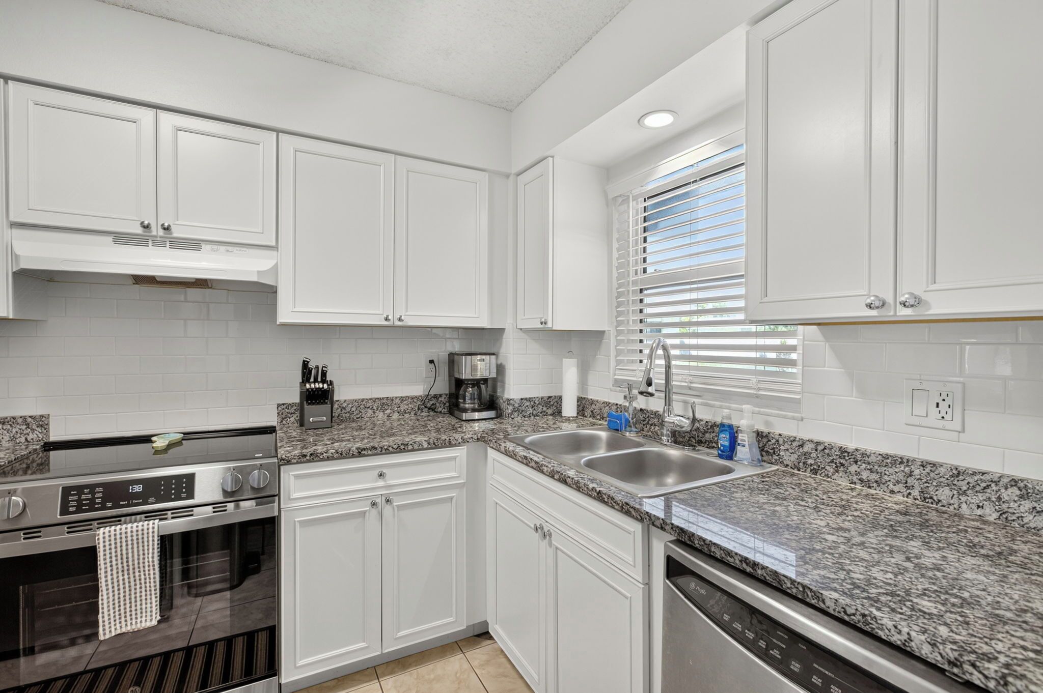 A modern kitchen with white cabinetry, granite countertops, stainless steel appliances, and a window with blinds.