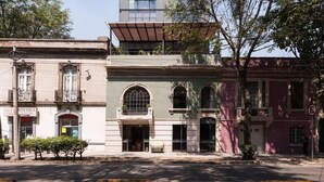 A row of historic buildings with arched windows and balconies, a central building with a flat roof, and a tree-lined street.