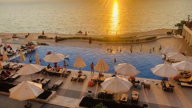 A resort pool area with sun loungers and umbrellas, surrounded by a landscaped deck and overlooking the sea. - Porto Dead Sea  (Sweimeh)