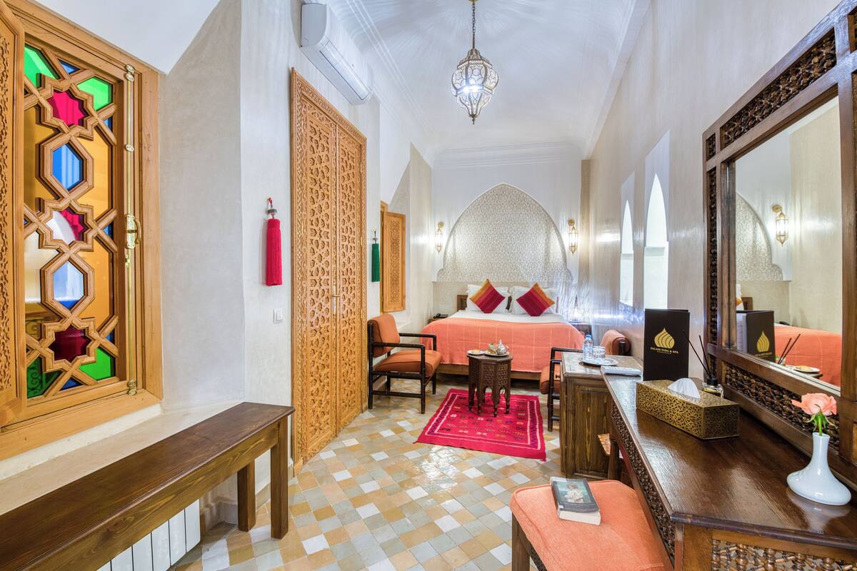 Una habitación de hotel con piso a cuadros, una cama con almohadas rojas y rosadas, un banco de madera, una mesita con un florero y un espejo grande.