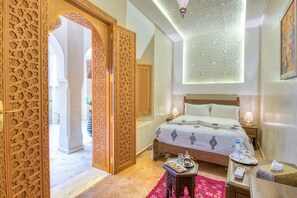 Quarto de hotel com uma cama grande, uma mesa de cabeceira de madeira, um tapete vermelho e portas de madeira detalhadas.