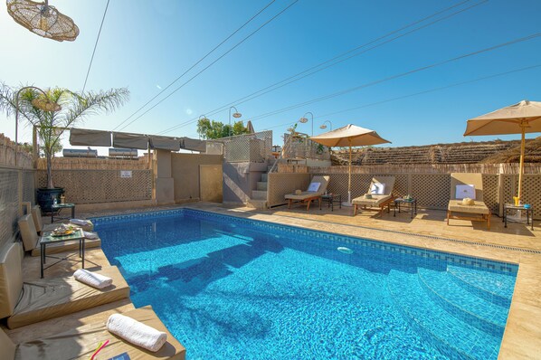 Une piscine d’un bleu éclatant, bordée de chaises longues et d’ombrelles, entourée d’une haute clôture et ombragée par des pergolas.