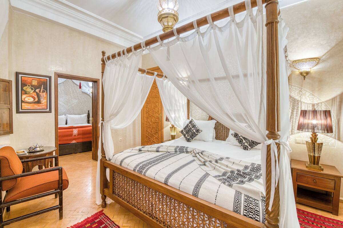 Una cama con dosel, un mesita de noche de madera con lámpara, una silla de mimbre y un tapete rojo estampado.