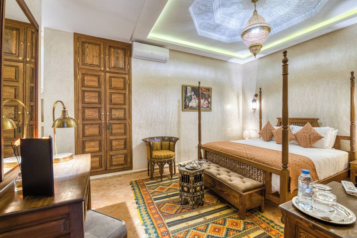 Una cama con dosel tradicional, un cabecero de madera, una mesita redonda con bandeja y una alfombra estampada.