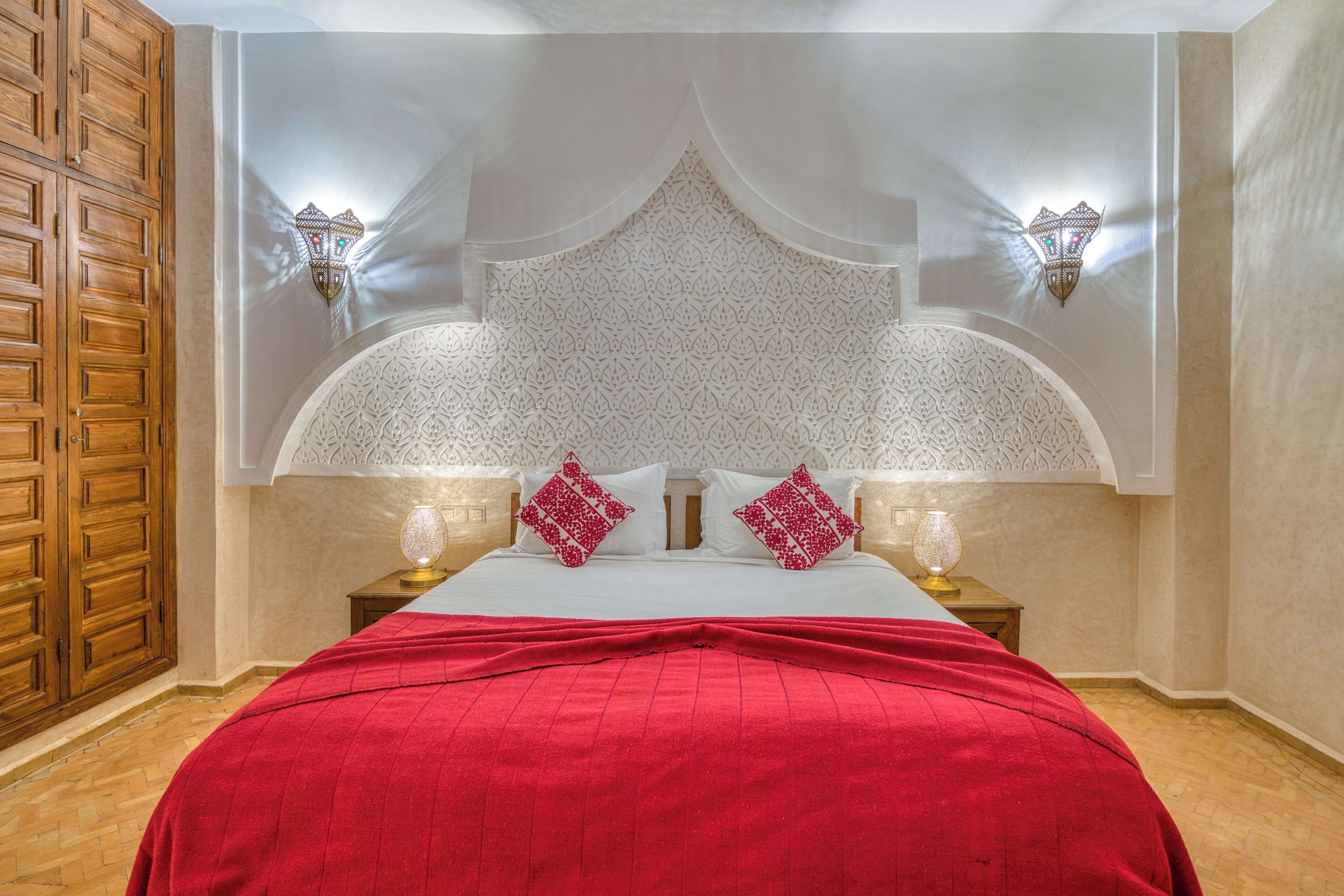 A bedroom with a large bed, ornate headboard, two wall sconces, and a wooden wardrobe.