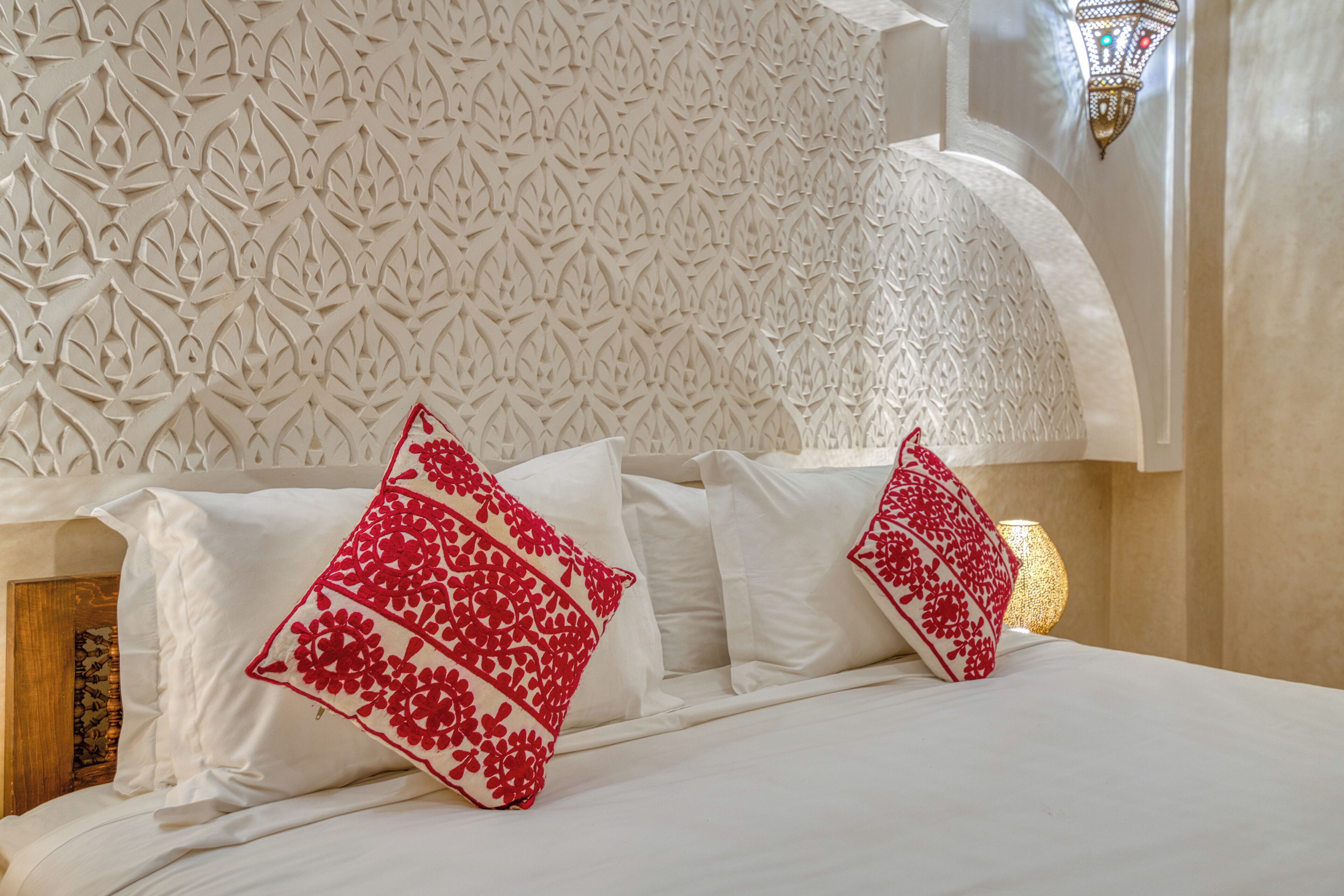 Een bed met witte lakens en rode kussens met bloemen, een decoratieve wand en een wandlamp.