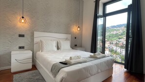 A modern bedroom with a large bed, two bedside tables, and a view of a cityscape through a large window. - Sea and Sun 4 You - Villa Oliveira (Funchal)