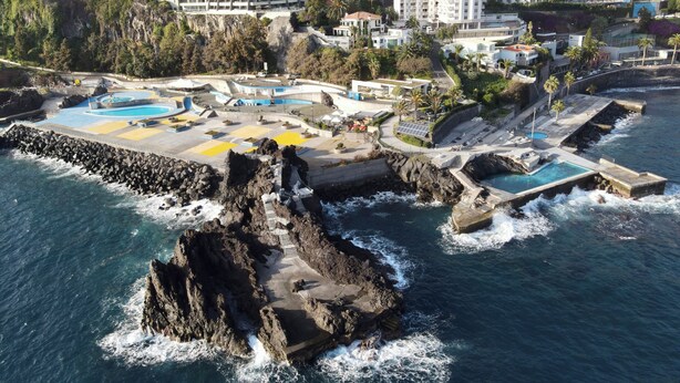 An aerial view of a coastal resort with multiple swimming pools, a rocky shoreline, and residential buildings in the background. - Sea and Sun 4 You - Villa Oliveira (Funchal)