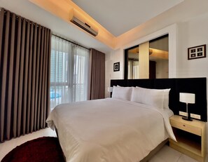 A hotel room with a large bed, a bedside table with a lamp, a window with curtains, and an air conditioning unit.