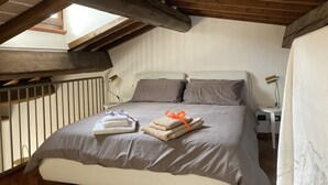 A bedroom with a sloped ceiling, wooden beams, a bed with grey bedding, two bedside tables with lamps, and towels on the bed. - Loft Albinelli in the heart of Modena (Modena)