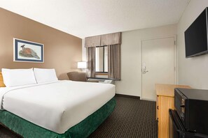 Een hotelkamer met een bed, een televisie, een bureau, een lamp, een raam met gordijnen en een ingelijst schilderij aan de muur.