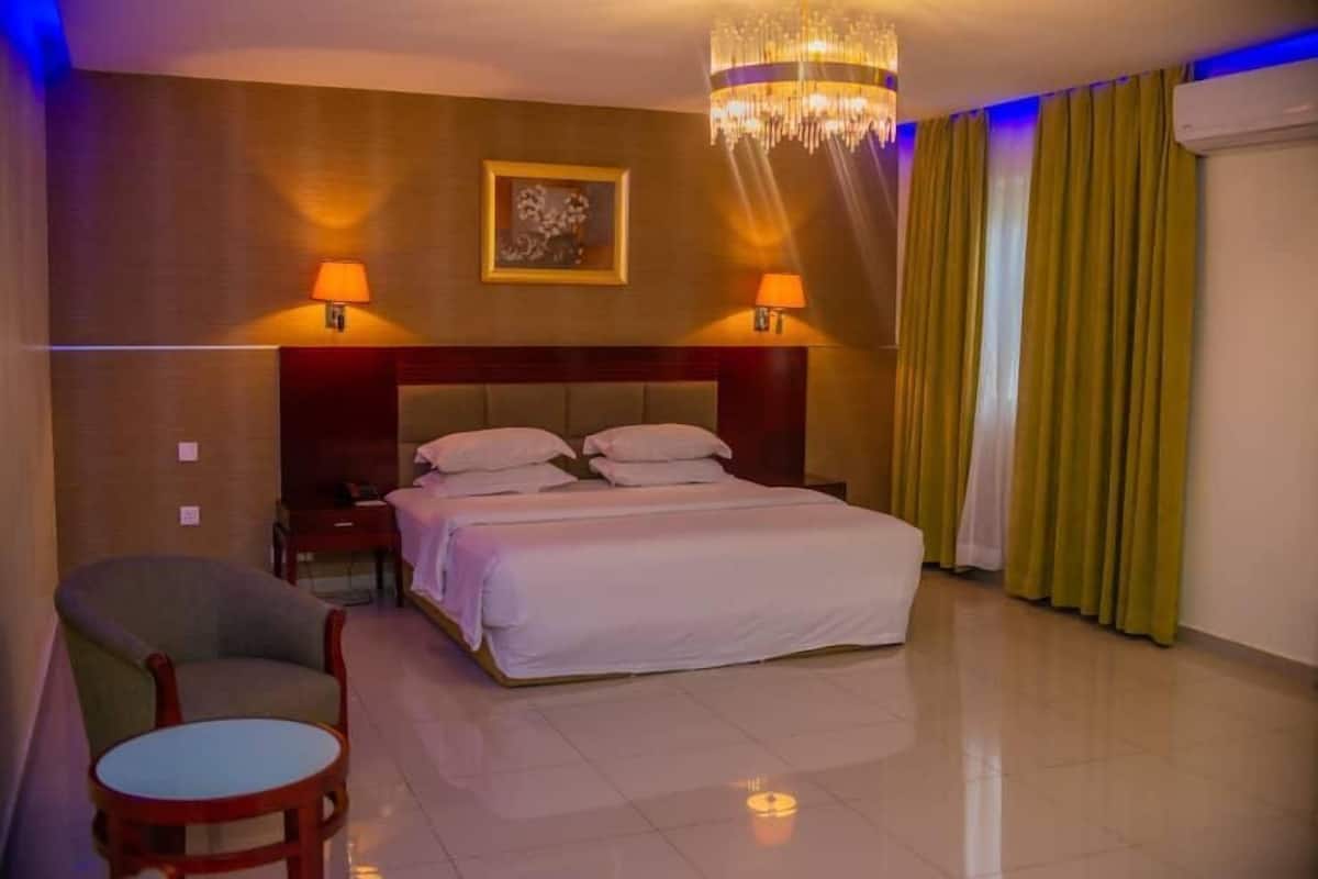 A hotel room with a bed, two bedside lamps, a chair, a small table, a chandelier, and a painting on the wall.