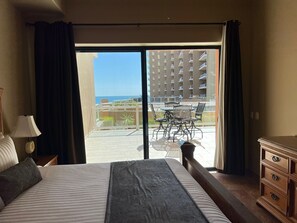 A hotel room with a bed, a dresser, a lamp, and a view of a balcony with outdoor furniture and a building in the background.