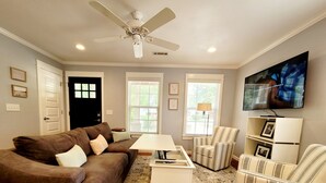 Ein Wohnzimmer mit braunem Sofa, gestreiften Sesseln, einem weißen Couchtisch, einem Deckenventilator und einem an der Wand montierten Fernseher.