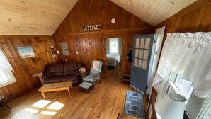 A cozy cabin interior with a wooden ceiling, a sign reading "COTTAGE", a brown leather sofa, a wooden armchair, a small table, and a window with curtains.