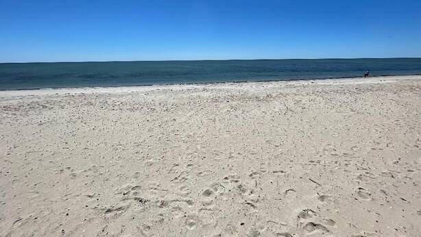 A wide sandy beach with footprints leading to the water, a clear blue sky, and a distant person walking.
