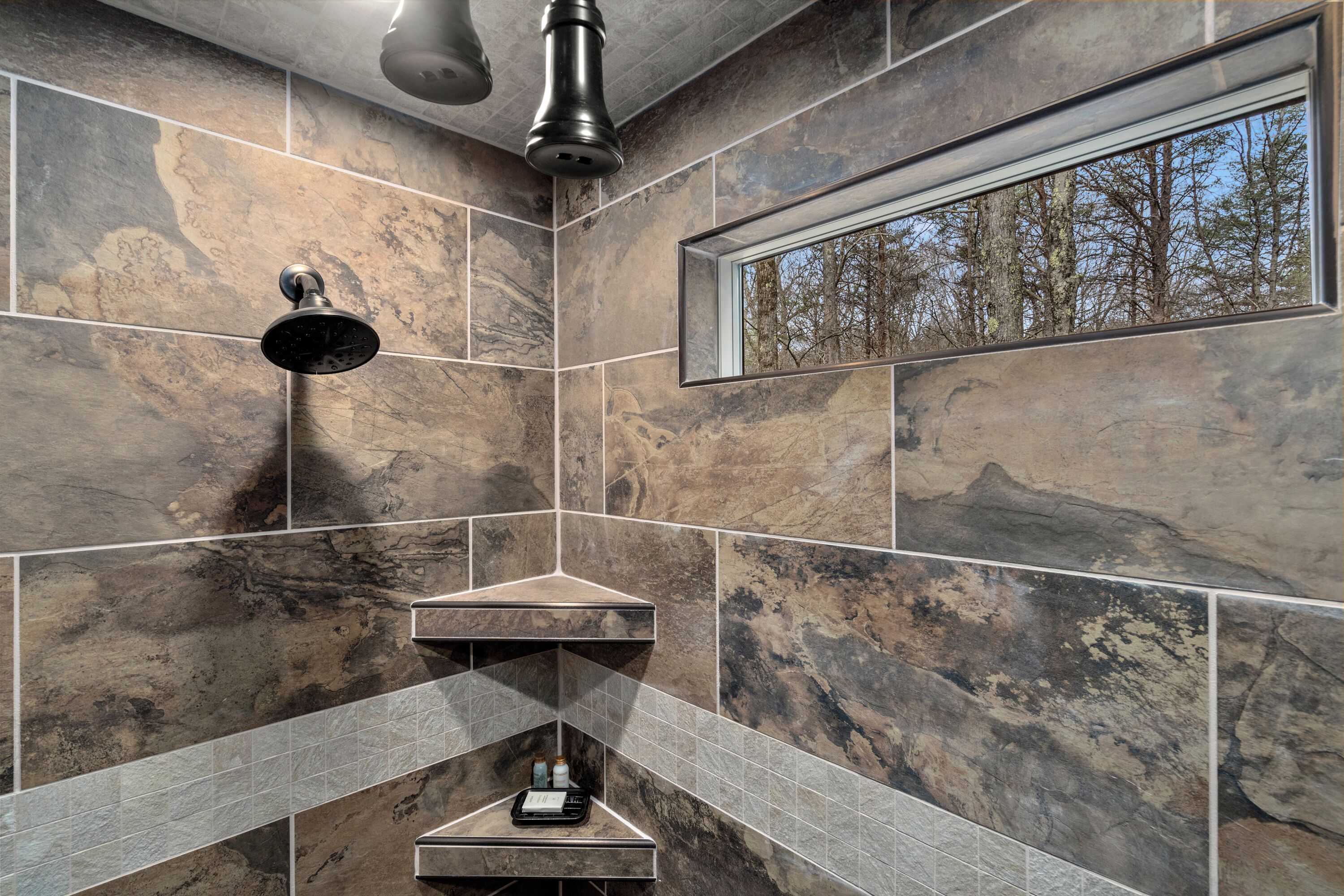 A modern bathroom with a shower, a window with a forest view, and a shelf with toiletries.