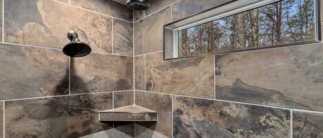 A modern bathroom with a shower, a window with a forest view, and a shelf with toiletries.
