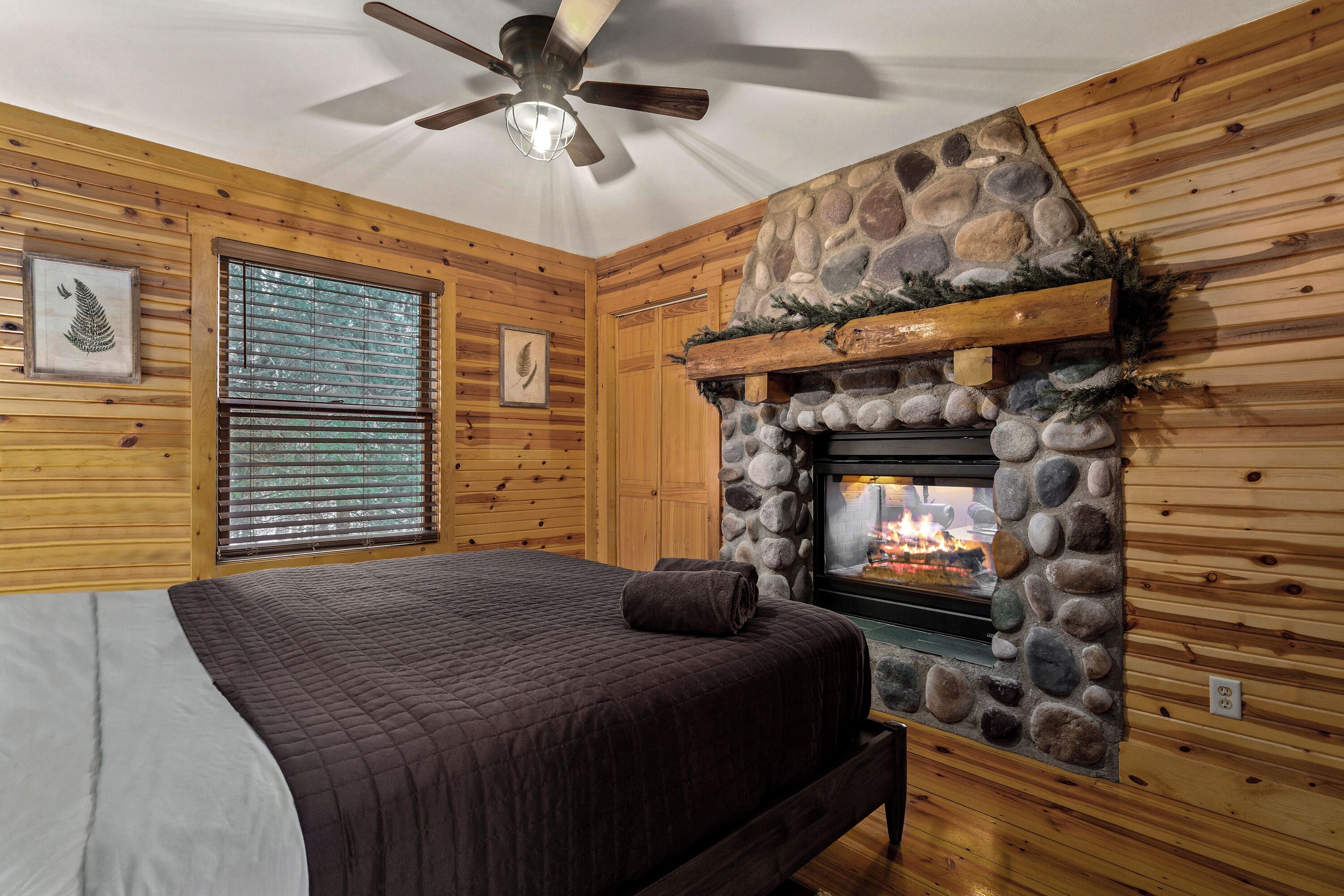 A bedroom with a stone fireplace, wooden walls, a bed with a quilt, and a ceiling fan.