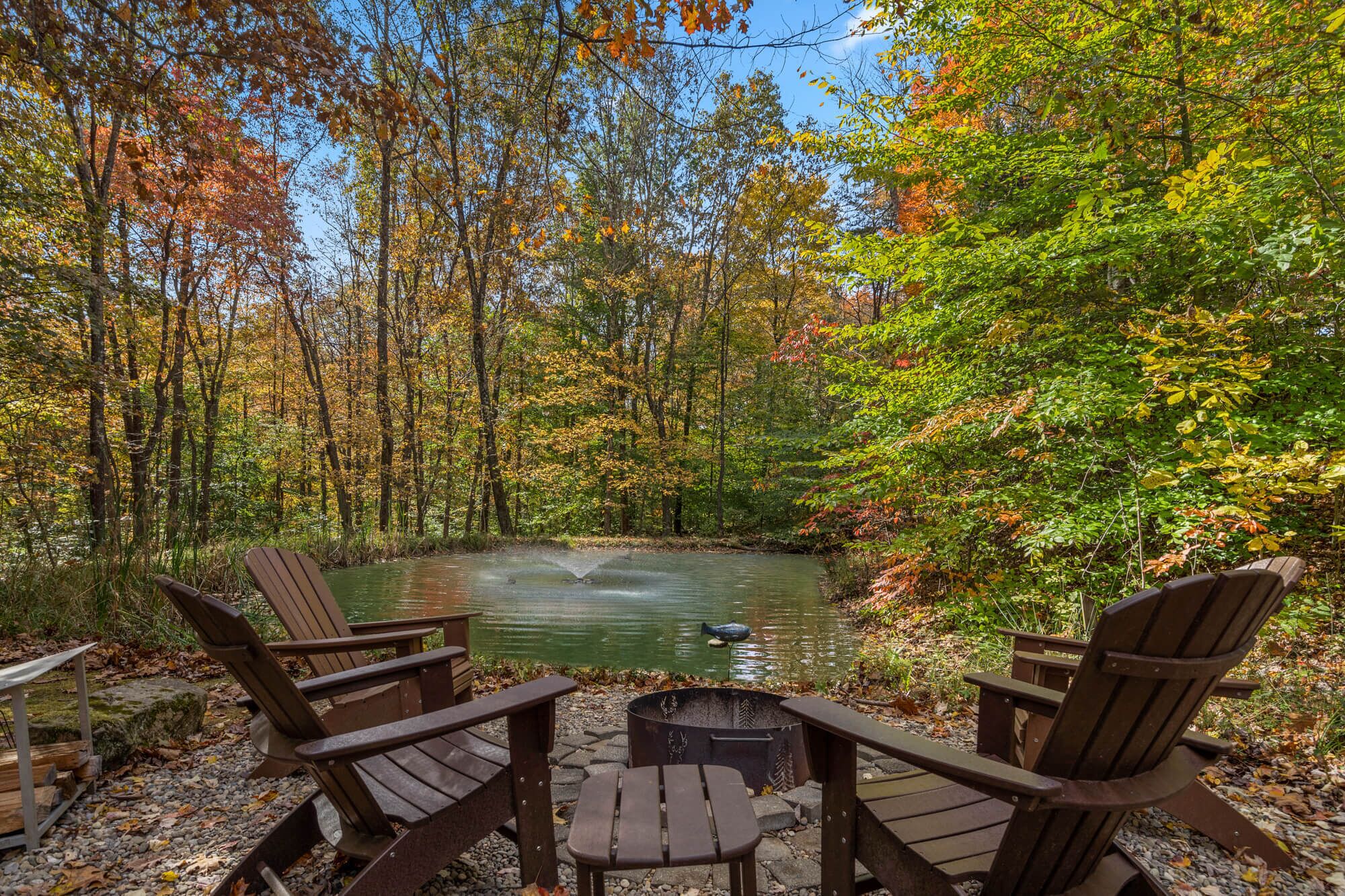 Un espacio al aire libre tranquilo con sillas Adirondack y una chimenea, rodeado por un bosque de follaje otoñal.