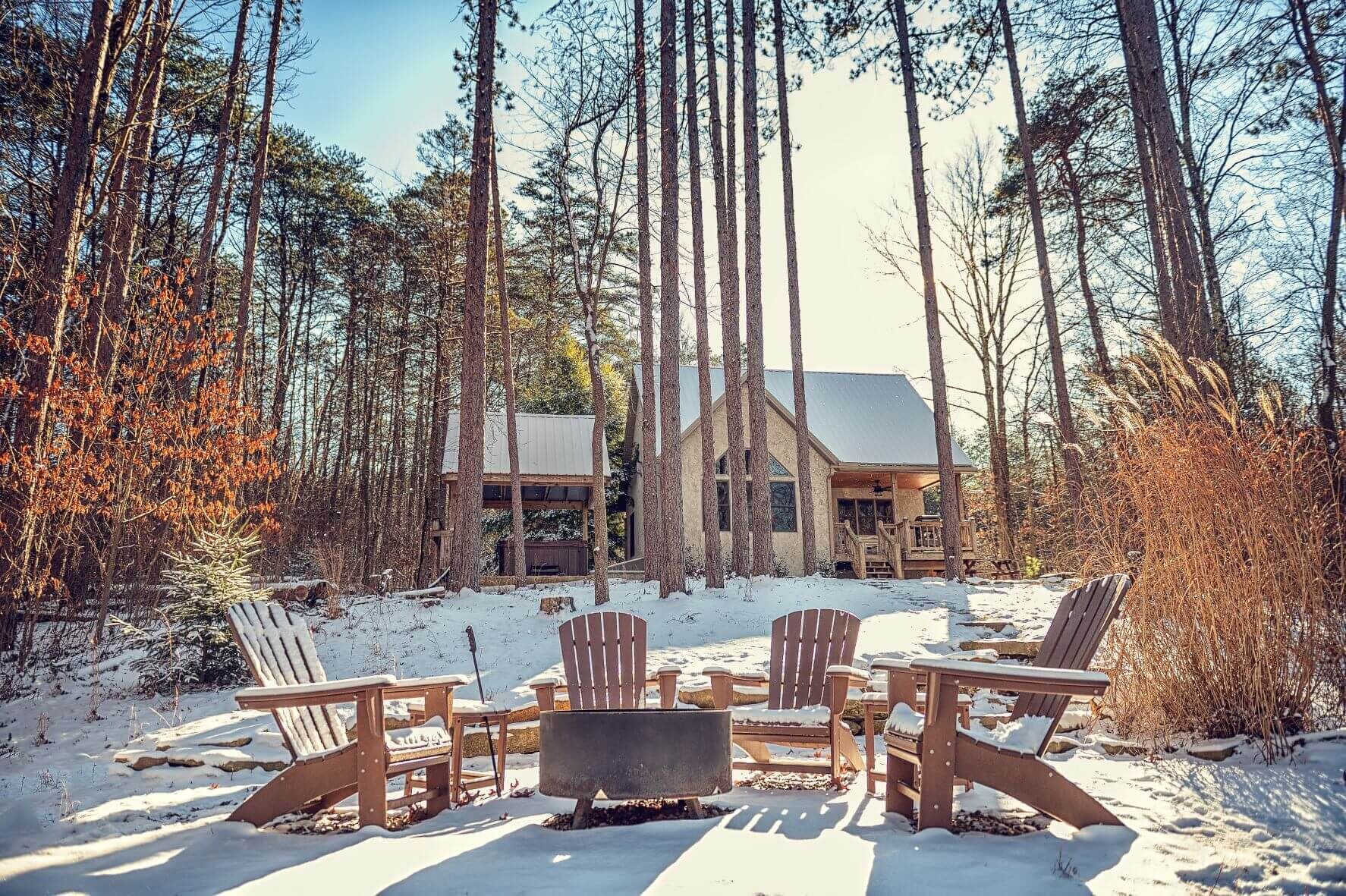 A cabin surrounded by tall trees, with a snow-covered deck and wooden chairs.