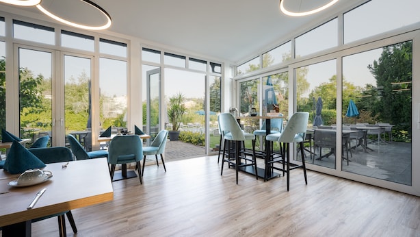 A modern dining area with large windows, light wood flooring, and teal chairs. - Hotel Villa Flora (Velden am Wörther See)