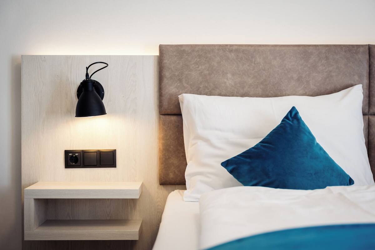 Een net opgemaakt bed met een wit en blauw kussen, een houten nachtkastje met een zwarte lamp en een schakelaar aan de muur.