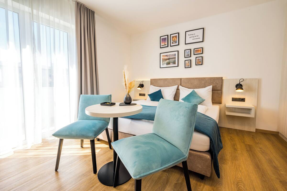 Een moderne hotelkamer met een bed, twee blauwe stoelen, een klein rond tafeltje en een nachtkastje met een lamp.