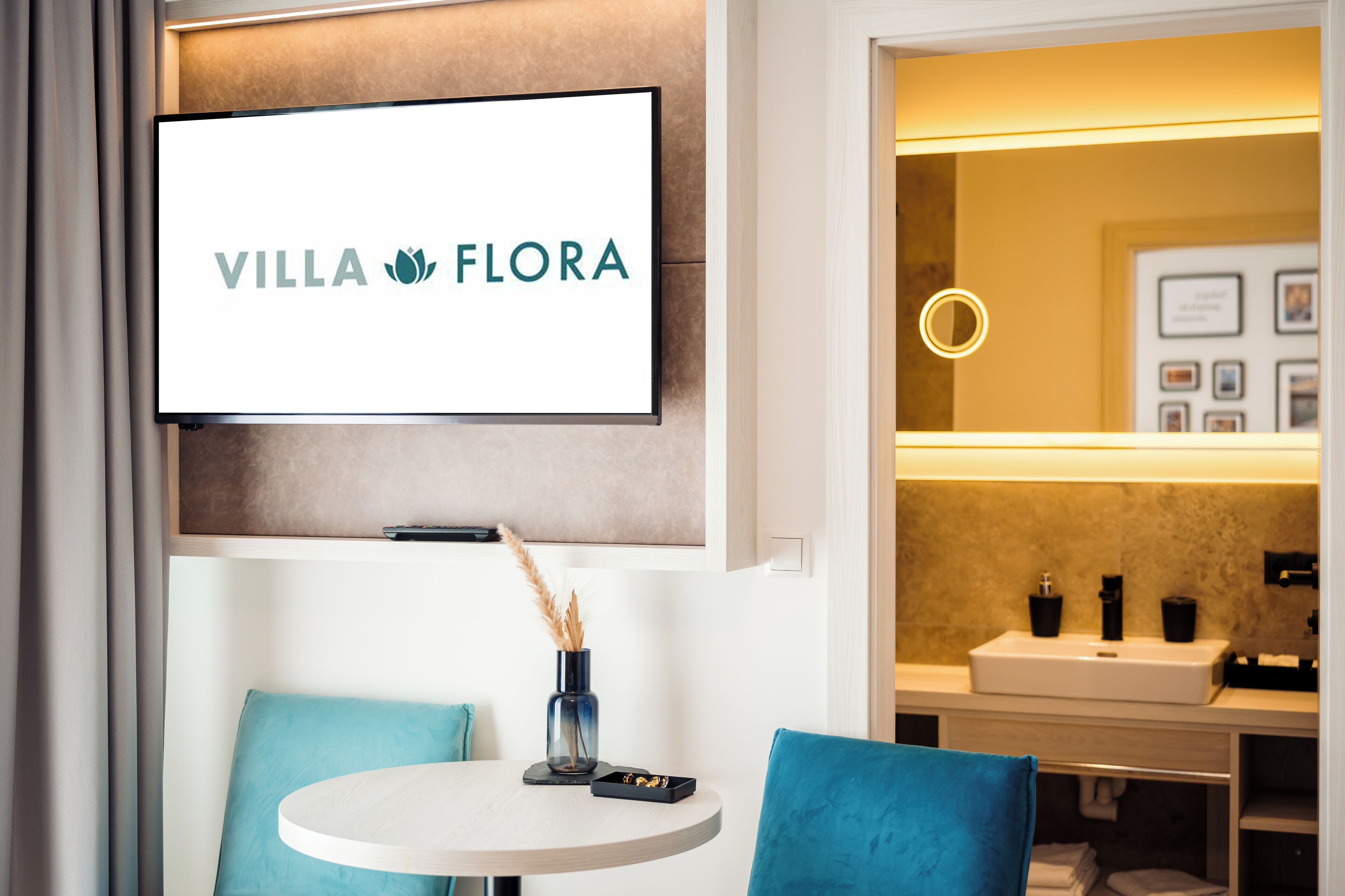 A modern hotel room with a mounted television displaying 'VILLA FLORA', a round table with a vase and remote control, and a bathroom with a mirror and sink.