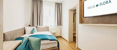 A hotel room with a bed, a desk, a TV mounted on the wall, and a window with curtains.