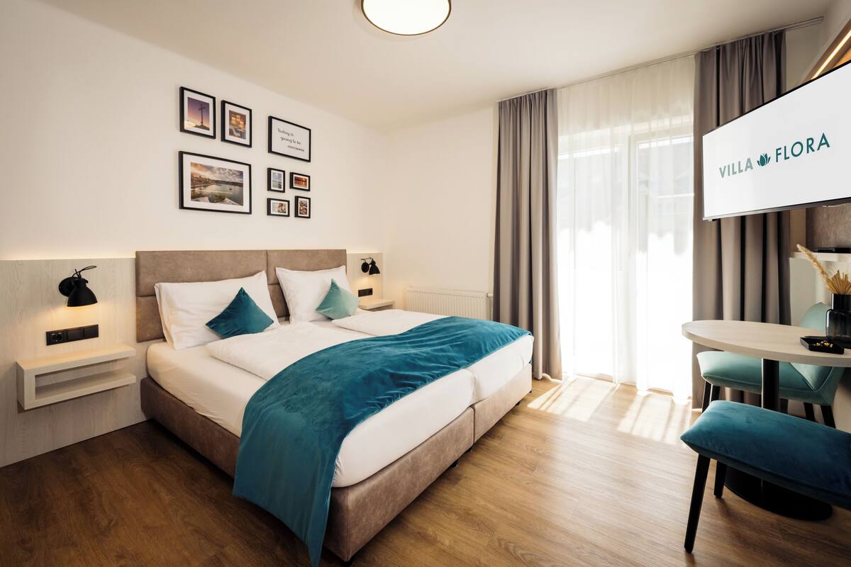 Een moderne hotelkamer met een groot bed, nachtkastjes, een bureau en een televisie waarop 'VILLA FLORA' staat.