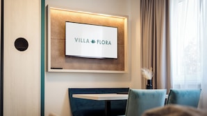 A hotel room with a mounted TV displaying "VILLA FLORA", a wooden wall, a window with curtains, and a seating area with a table and chairs. - Hotel Villa Flora (Velden am Wörther See)