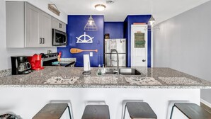 A modern kitchen with white cabinets, a granite countertop, a coffee maker, and a bar area with stools. - Beach, Please! (Carolina Beach)