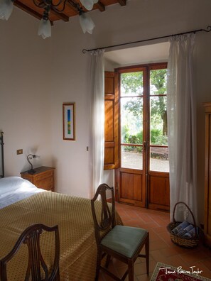 Ein Schlafzimmer mit Bett, Stuhl, Schreibtisch mit Lampe, Fenster mit Vorhängen und Blick ins Grüne.