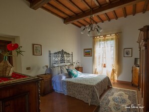 Quarto com cama, cabeceira de madeira, candelabro, mesa de cabeceira, cômoda e janela com cortinas.