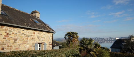 Un edificio de piedra con una ventana pequeña y una puerta, rodeado de vegetación y vistas a un cuerpo de agua y edificios a la distancia.