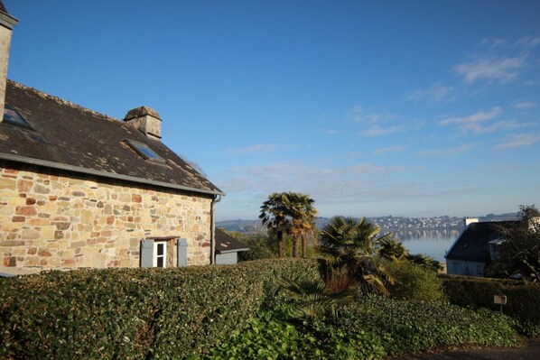 A stone building with a small window and a door, surrounded by greenery and a view of a body of water and distant buildings. - La Cidrerie du port (landevennec)