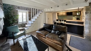 A modern living room with a leather sofa, a glass coffee table, and a stone wall. A staircase leads to a kitchen area with a bar.