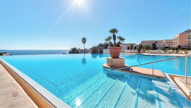 A clear blue infinity pool with a view of the sea, surrounded by palm trees and a building with balconies.